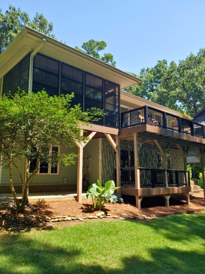 A multi-level outdoor living space featuring a screened-in porch on the upper level and a deck on the lower level, connected to a light green sided house. A lush green lawn and trees are in the background.